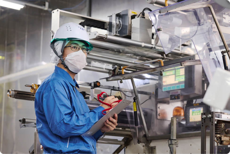 生産設備の点検を行う作業員。ヘルメットとマスクを着用し、クリップボードを手に持ちながら機械の状態を確認している。