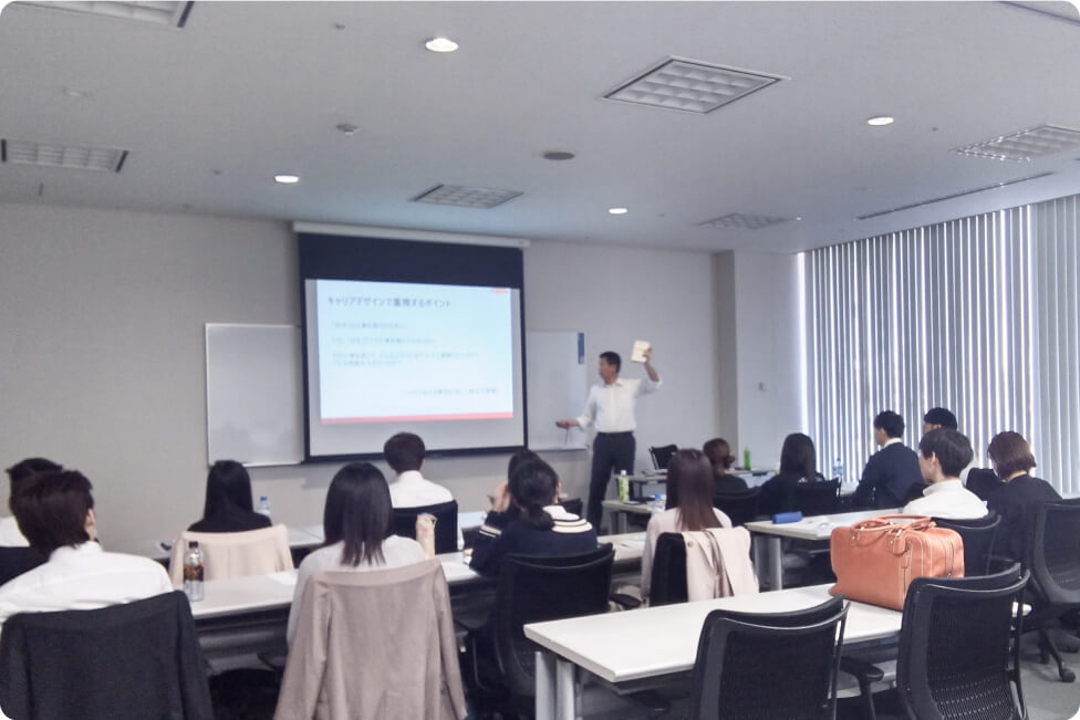 会議室で実施されている研修の様子。講師がスクリーンを使って説明し、受講者が資料を見ながら話を聞いている。